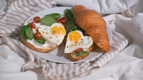 Valentine's day concept. Woman eating breakfast in bed on Valentine's Day. Sandwiches with croissant, bagel, cream cheese and fried eggs hearts on white bed.