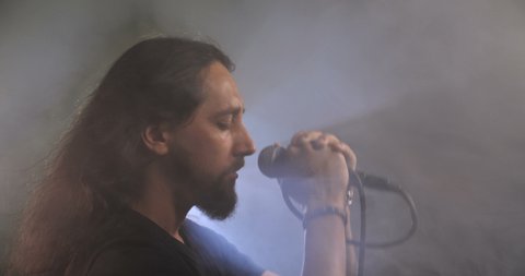 Dramatic close-up portrait of a long-haired Caucasian male, brunet with a beard against a black background with smoke, wind and backlight. Rock vocalist screaming to microphone. 50 fps 4k