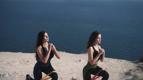 Slow motion two beautiful young brunette in sportswear doing squats with an elastic band on the background of the sea on sunny morning. Concept sports tour to exotic countries