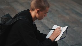 Portrait of handsome student guy looking dreamy making notes in notebook studying on stairs outdoor