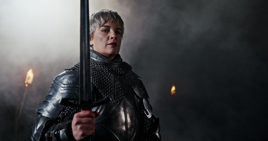 Determined medieval woman in armor with sword. Brave and powerful woman wearing warrior armor and holding sword representing Joan of Arc standing in dark with flames burning
