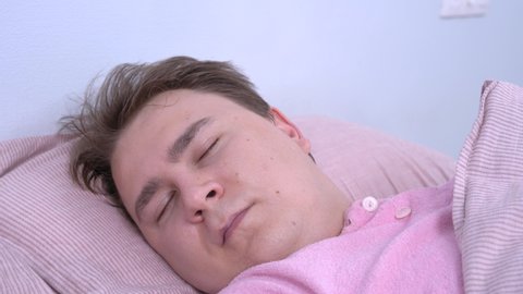 Young caucasian man with somnambulism in pink pajamas sleeps sweetly at home, mumbles in his sleep and smiles because he sees pleasant dream, close up.