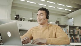 Smiling employee in headphones using laptop, looking at screen,mixed race man making video call , distance learning language concept, call center operator working