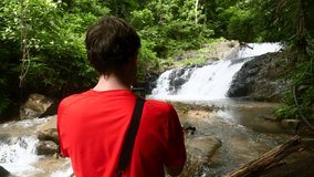 Back View of Traveler Man in Red T-shirt Enjoying Beautiful Waterfall Using Mobile Phone and Shooting Photo or Video for Social Network in Rainforest. Great Wild Jungle Nature, Lake and Forest