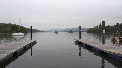 A small sailing yacht sails toward a jetty in a marina on the shores of Lake Windermere in the Lake District, England, UK