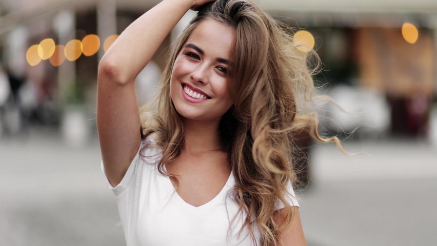 Beautiful fashionable model smiles, posing outdoors and straightens her long hair. Charming blonde look in slow motion. Portrait of gorgeous girl looking to camera