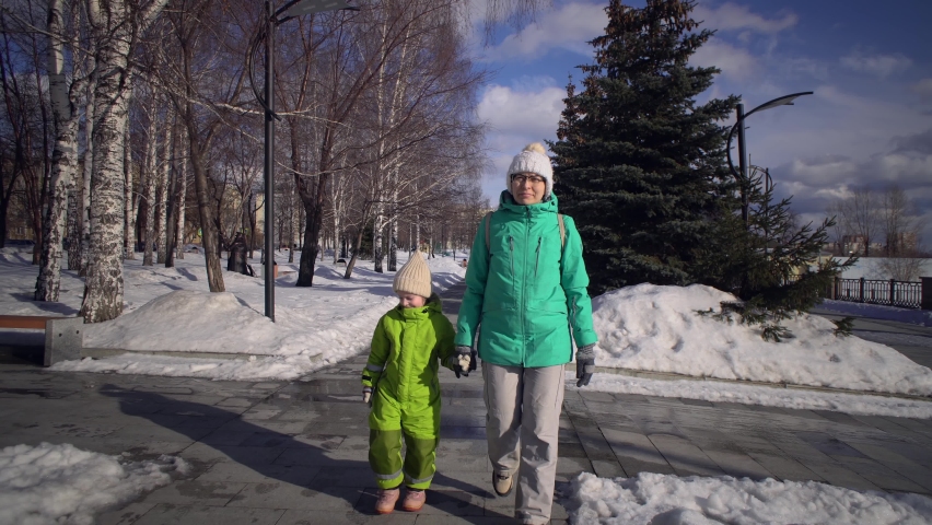 Young mother and cute happy child hold hands as they walk along the sidewalk next to snowdrifts in winter park.