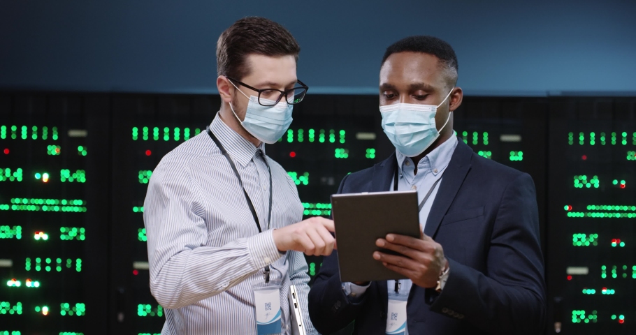 Close up portrait of mixed-races male security analytics in medical masks typing on tablet and speaking standing in datacenter. Coworkers discussing technical issue in server room. Big data protection