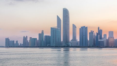Abu Dhabi city skyline with skyscrapers before sunrise with water reflection night to day transition timelapse from the breakwater near cultural village. Few clouds on morning sky