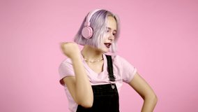 Pretty girl with dyed violet hair listening to music, smiling, dancing in headphones in studio against pink background. Music, dance, radio concept, slow motion.