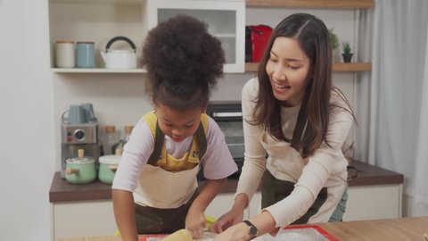 Lovely African daughter rolling out dough by using a rolling pin on wooden table while smiling beautiful Asian mother standing to watch and teaching next to her while making dough together in kitchen.
