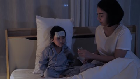 Mother caring for sick daughter lying in bed with Antipyretic gel sheet on the forehead and giving eat liquid cold medicine. Medical and child health concept in night shots. 