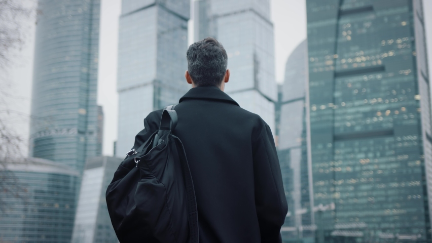 Man in black coat going towards business city skyscrapers in the evening, lights in the windows. Gimbal shot of young businessman, no face, with bag walking to business buildings Royalty-Free Stock Footage #1063617604