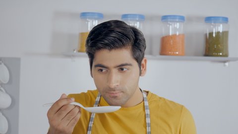 Smart and smiling student tasting his food - home alone cooking. Closeup shot of a modern Indian male checking the food with a small ladle in his kitchen - modern cooking