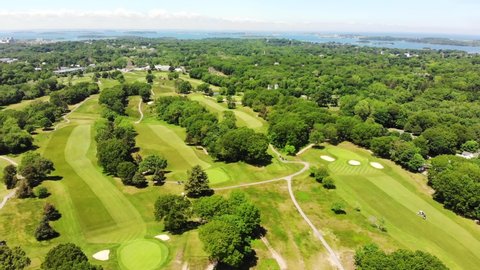 A daytime aerial view of a golf course as the drone flies toward the harbor or bay showing islands, golfers dot the green grass below and trees dot the landscape.