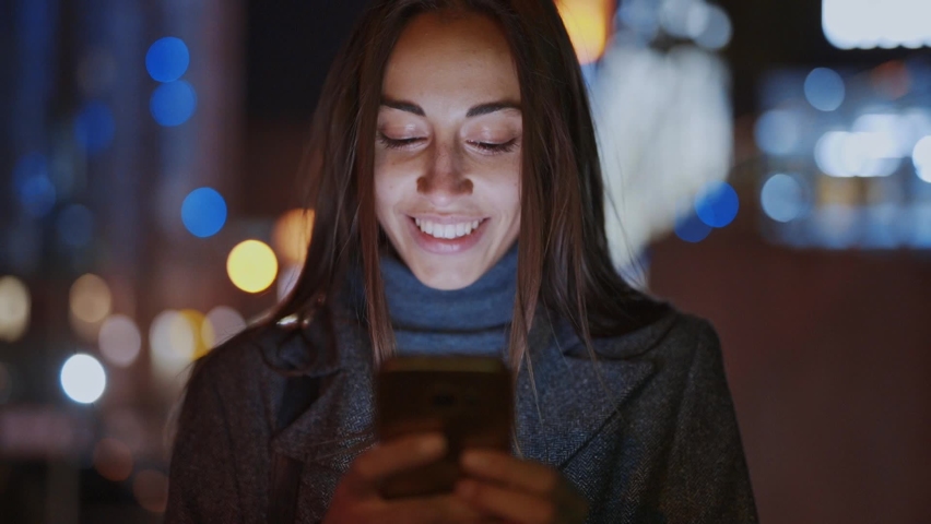 Closeup of prety smiling woman using smartphone walking at night city, slowmotion fullHD stock footage, handheld camera shot, clip has noise