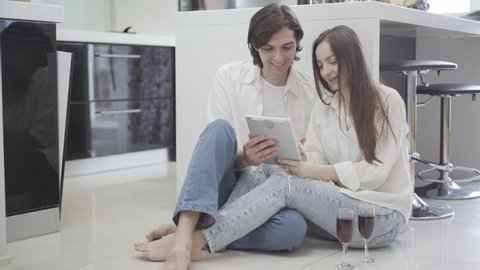 Happy relaxed young couple sitting on kitchen floor with wine glasses and using tablet. Portrait of positive Caucasian man and woman choosing online goods for new appartment. Rental and moving.