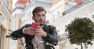 Bike messenger with backpack looks photos in his smartphone. Young man with bicycle stands on the street, uses smartphone and smiles. Cinema 4K 60fps video