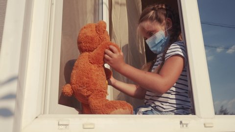 bored kid in medical mask in home quarantine coronavirus sitting by covid 19 the window. child with a toy teddy bear in protective mask looking out the window. coronavirus epidemic prevention concept