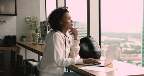 Pensive serious african 35s freelancer woman sit at table in modern office room alone working using laptop looking out the window thinking over business issue solution make telecommute job from home