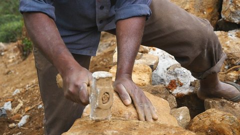 Close up of male hands of indian stone worker hits a stone with sledgehammer for construction flowerbed. Arms of stonemason cuts block of granite with a hammer in botanical garden. Slow motion