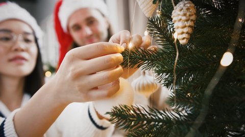 Young blurred mixed race couple, man and woman in Santa hats are smiling while decorating green Christmas tree with white bauble at home. Happy New Year, holiday. Close up, slow motion