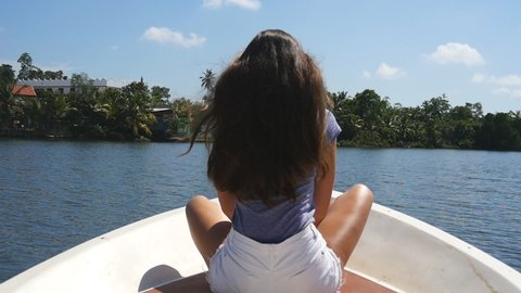Unrecognizable girl with blowing long hair sits on bow of boat and admires a beautiful nature landscape. Young woman rests on deck of yacht and enjoys summer travel. Concept of vacation or holiday