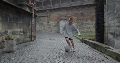 Front view of young kid dribbling at empty old europian city street. Talanted teenager boy running and kicking football ball. Concept of lifestyle, childhood, sport.