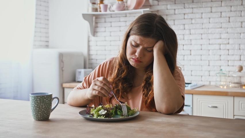 Healthy nutrition, eating disorder. Plus size woman sitting at table, feeling unhappy with diet not wanting to eat salad | Shutterstock HD Video #1063655665