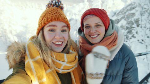 Young beautiful female friends looking at camera, smiling, telling about nature and sending air kiss while standing on mountain peak in national park on winter day and talking on video call