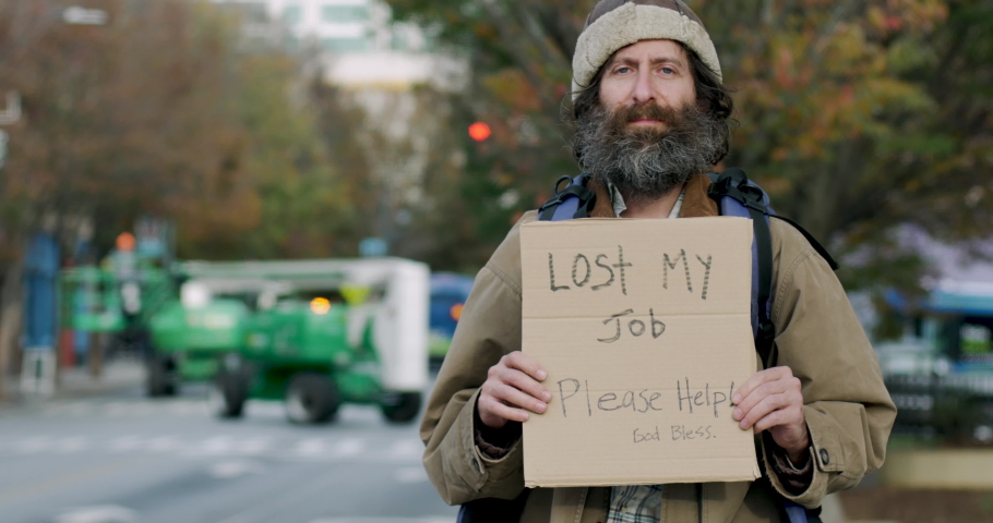 Portrait of an unemployed homeless looking man with a beard holding a sign asking for help looking directly at camera while standing on the side of a city street | Shutterstock HD Video #1063658956