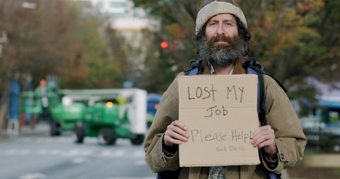 Portrait of an unemployed homeless looking man with a beard holding a sign asking for help looking directly at camera while standing on the side of a city street
