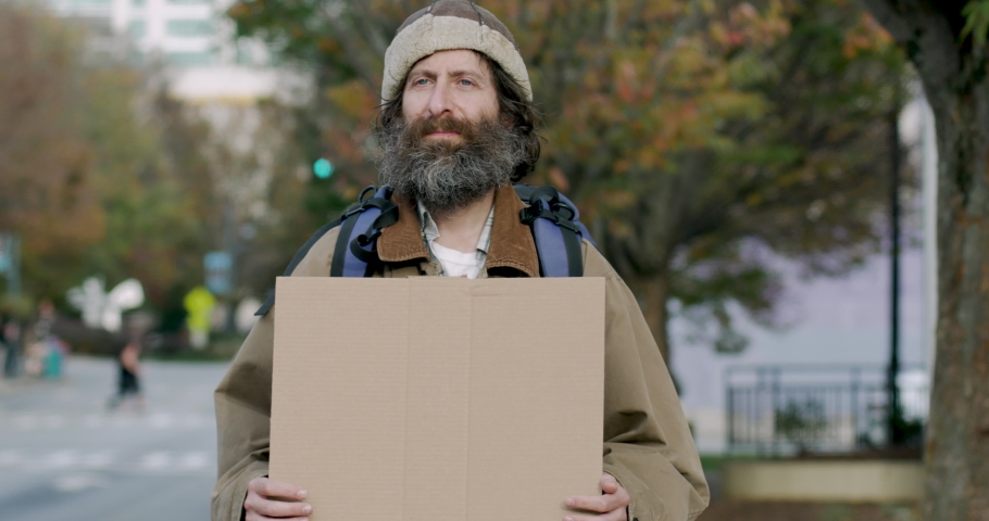 Homeless looking man with a beard wearing a hat, coat and backpack holding a blank cardboard sign standing in the city street