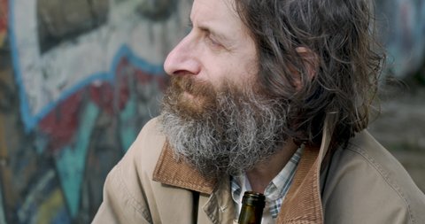 Alcoholic man in his 40s or 50s with a beard drinking out of a paper bag and wiping his face with the back of his hand while sitting outside