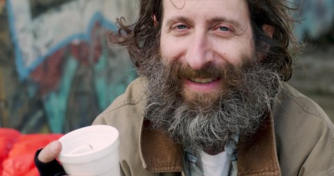 Portrait of a homeless Caucasian man in his 40s or 50s with a big beard smiling and laughing while looking at the camera