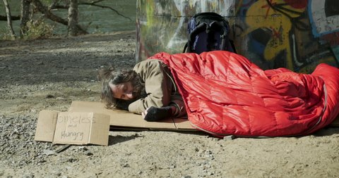 Homeless man lying on cardboard puts on his hat and tries to get comfortable under a sleeping bag
