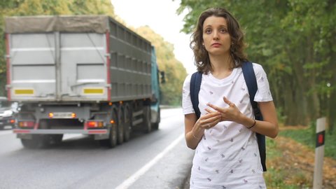 Brunette young woman with dark blue backpack is hitchhiking on the side of the road and a car is stopping by on trees background.