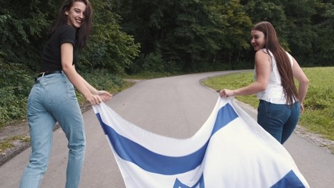 Two beautiful friends holding Israel national flag and going in a park. Independence Day.