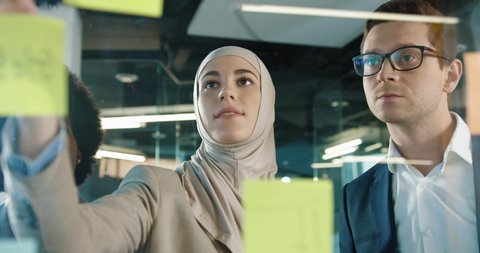 Close up of mixed-races male and female professional working in modern office. Beautiful Arabian female writing on glass board in cabinet and speaking with colleagues. Business concept