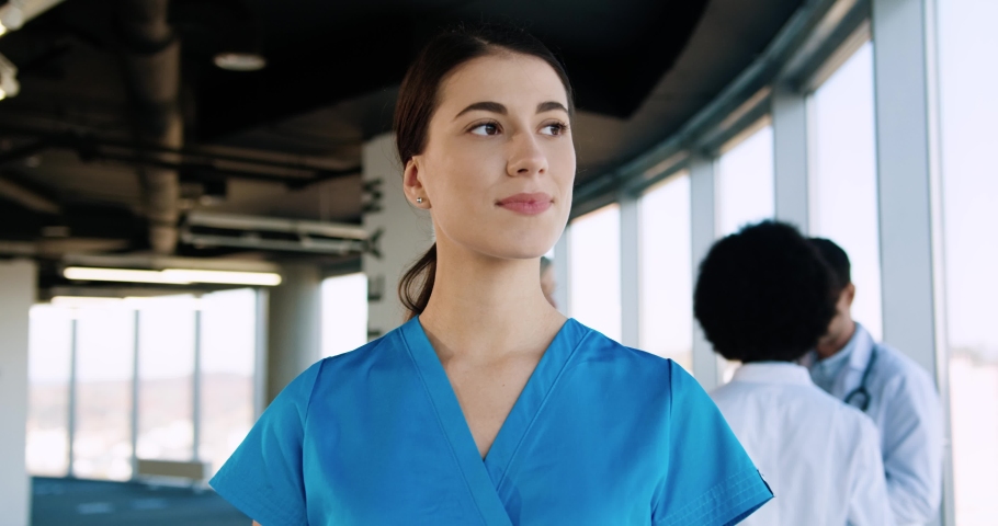 Close up portrait of cheerful young beautiful Caucasian female professional nurse healthcare assistant standing in modern hospital and smiling to camera in good mood. Doctors working on background | Shutterstock HD Video #1063665685