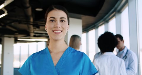 Close up portrait of cheerful young beautiful Caucasian female professional nurse healthcare assistant standing in modern hospital and smiling to camera in good mood. Doctors working on background