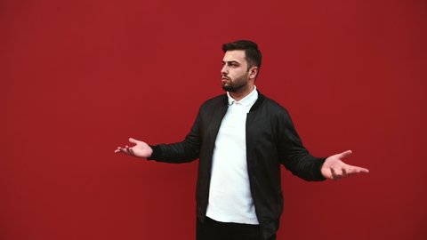 Confused bearded man in casual clothes stands against the background of a red wall and looks to the side with a puzzled face and spreads his arms.