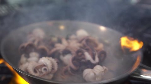 Preparing exotic fresh octopuses in metal frying pan with burning oil and thyme on stove orange flame in luxury seafood restaurant kitchen extreme close view.