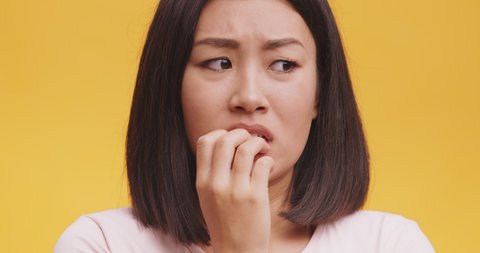 Nervous asian woman feeling panic, biting her nails, feeling worried and confused, orange studio background, close up portrait
