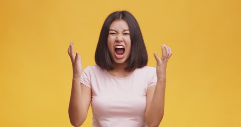 Emotional burnout. Young furious asian woman screaming loud and shaking hands, feeling rage and anger, orange studio background