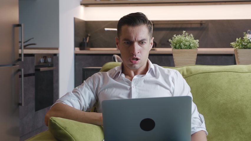 Angry male entrepreneur is experiencing anger and emotional stress while working on laptop while lying on sofa at home. A male freelancer feels angry and shows aggression while working at a computer Royalty-Free Stock Footage #1063688500