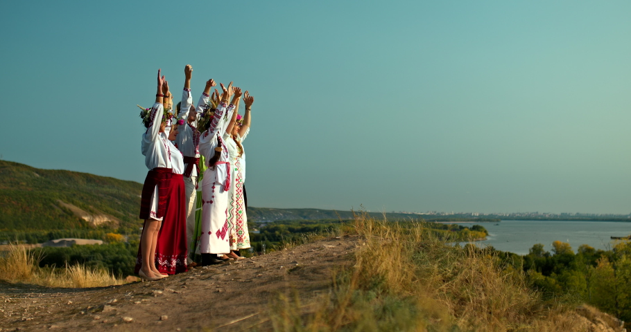 Group of women and men in folk costumes stand on the mountain and raise their hands up, folklore traditions and customs. Beautiful rural landscape. 4k, ProRes