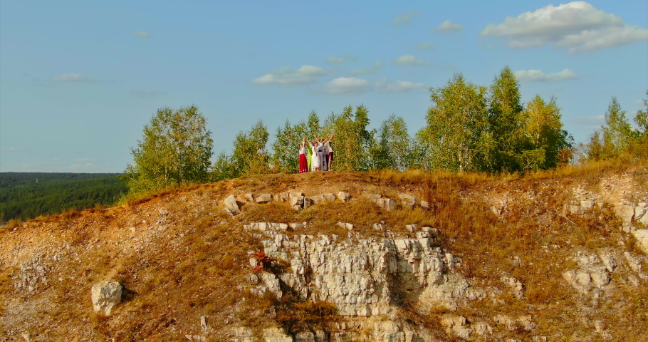 Aerial view, people in beautiful folk costumes and dresses stand on the hill and raise their hands up, folk ceremony. Slavic people stand in a round dance with their hands up, camera flight. 4k