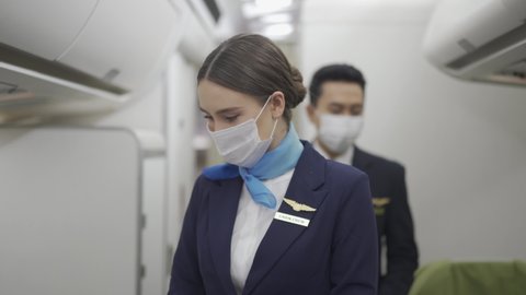 Caucasian female and Asian male flight attendants in a face mask for protective Covid-19 walk along aisle checking the passengers on safety standards with seat belt fastened before airplane take off.