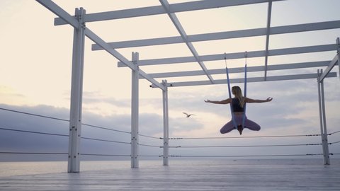 Young blonde woman in modern tracksuit does yoga exercises hanging on special slings fastened to wooden planks on sea empty pier against cloudy sky